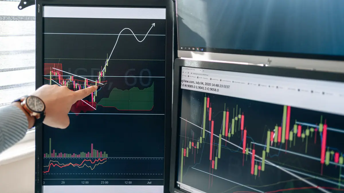 Person pointing at stock market graphs on dual monitors in a modern workspace.