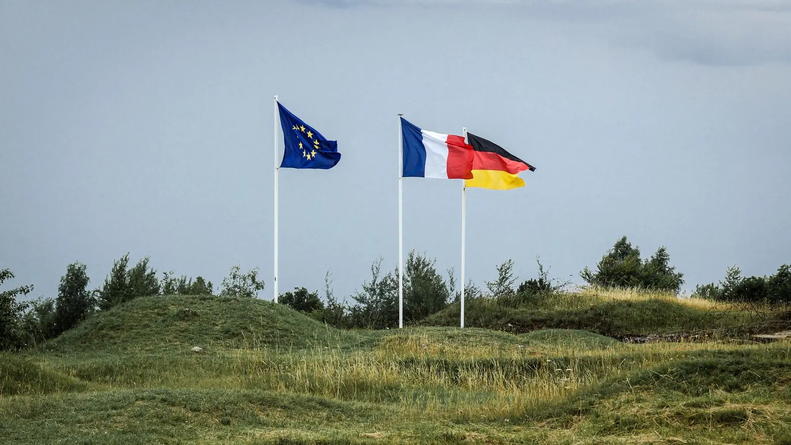 Flags of the European Union, France, and Germany waving on poles against a grassy hill and sky background.