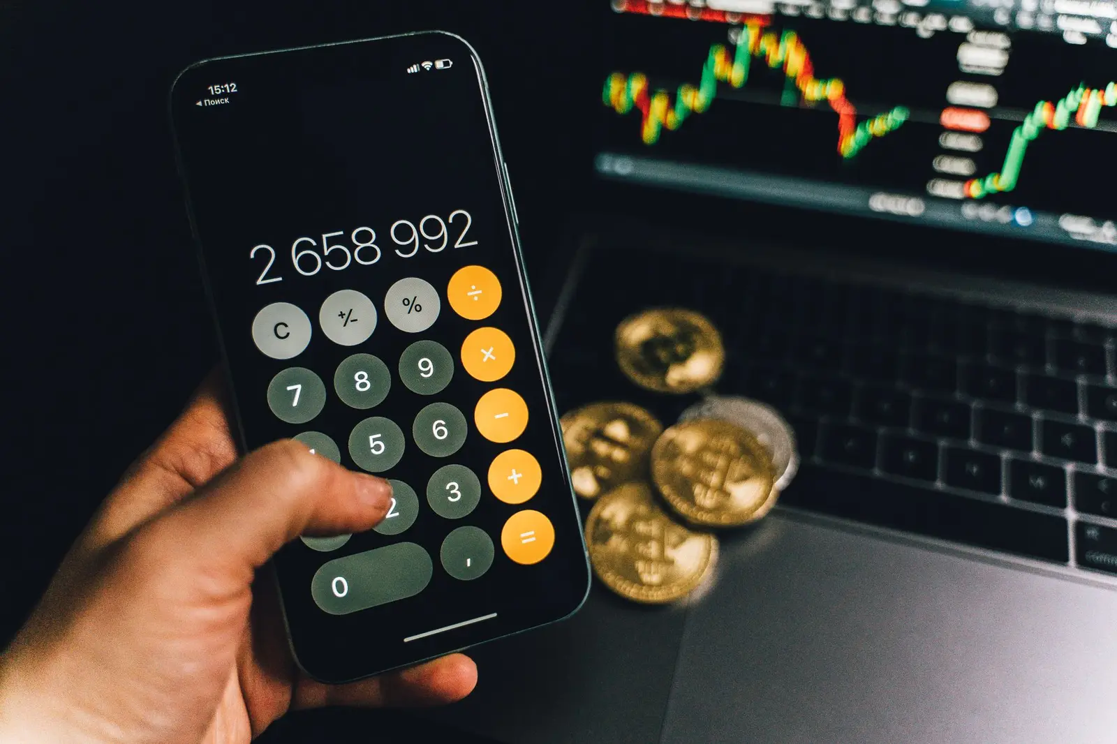 A smartphone displays a calculator app beside bitcoins and a laptop with a financial chart.